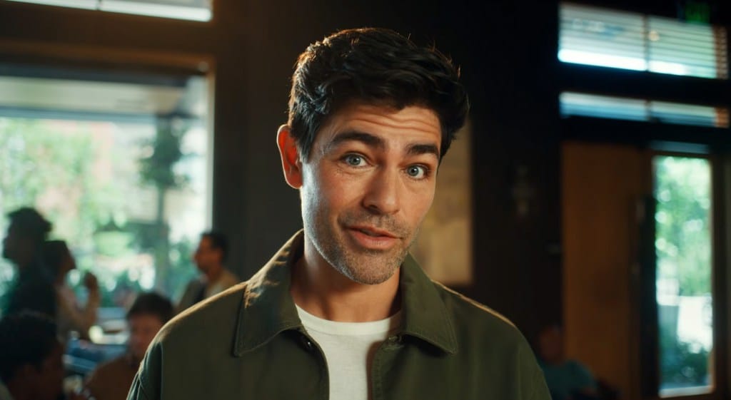 Adrien Grenier in a green jacket with a white shirt underneath, speaking directly to the camera in an ad.