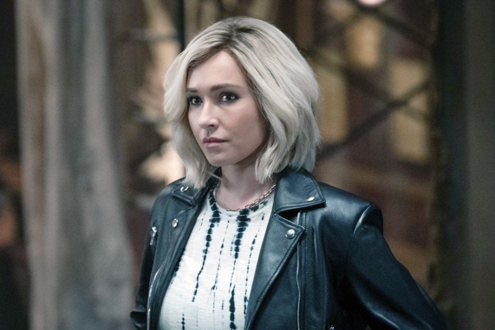 Person with short hair in a leather jacket stands confidently, looking to the side. Background is blurred, focusing on the person