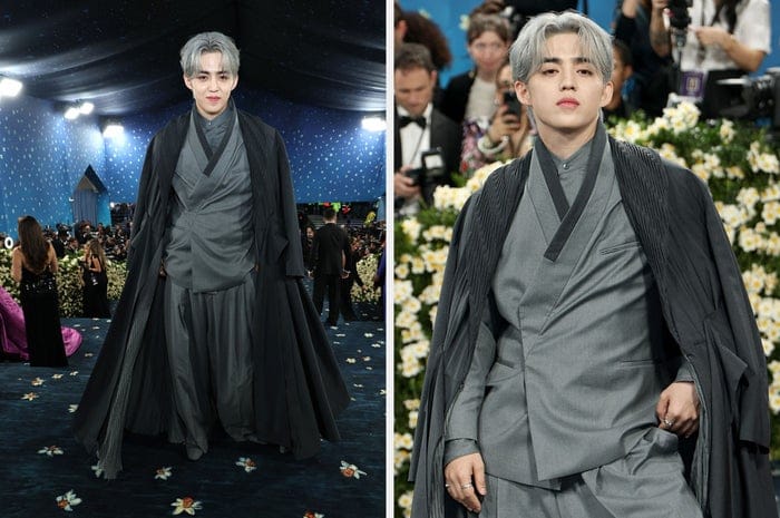 A person on a red carpet wears a flowing, layered gray outfit with a long cape, exuding a dramatic and elegant style
