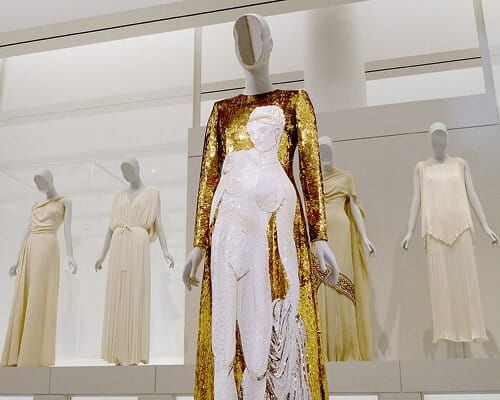 A gallery installation shot showing a historical garment displayed next to a painting or sculpture, emphasizing the pairing strategy and spatial relationship between the two mediums. Ideal...