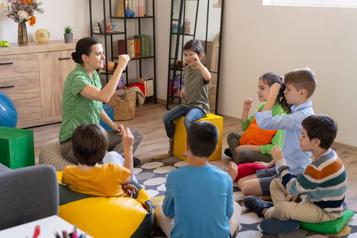 Child practising sign language while others watch as a teacher facilitates the teaching session, sitting in a circle on the floor 