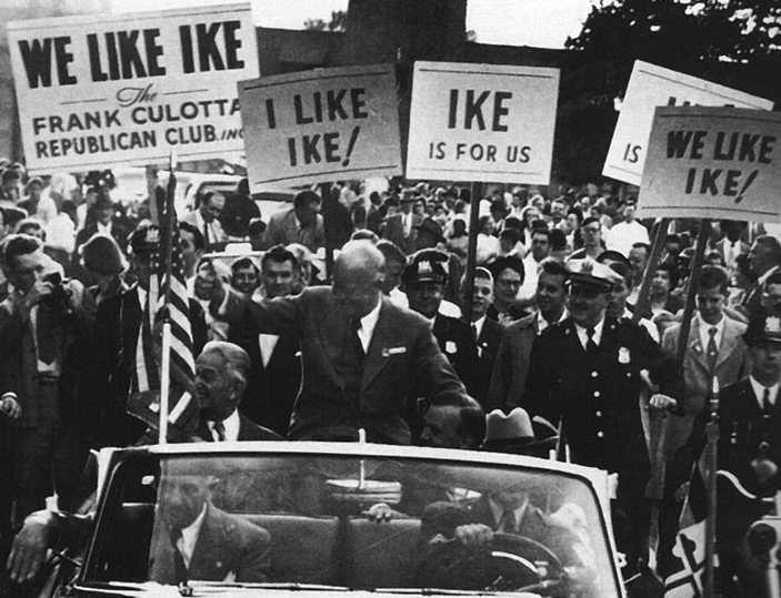 US president Dwight “Ike” Eisenhower in a motorcade with supporters holding up placards.