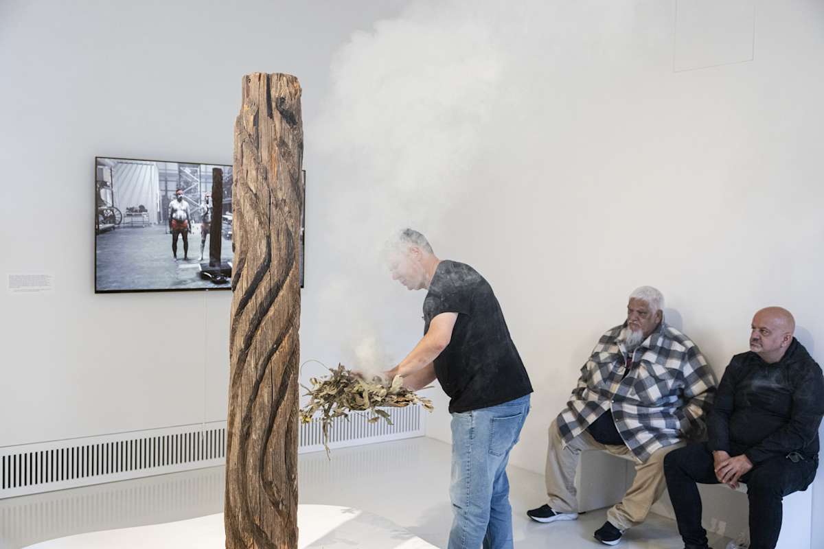 Two men sit watching another man perform an Indigenous smoking ceremony with a sacred tree in a romm