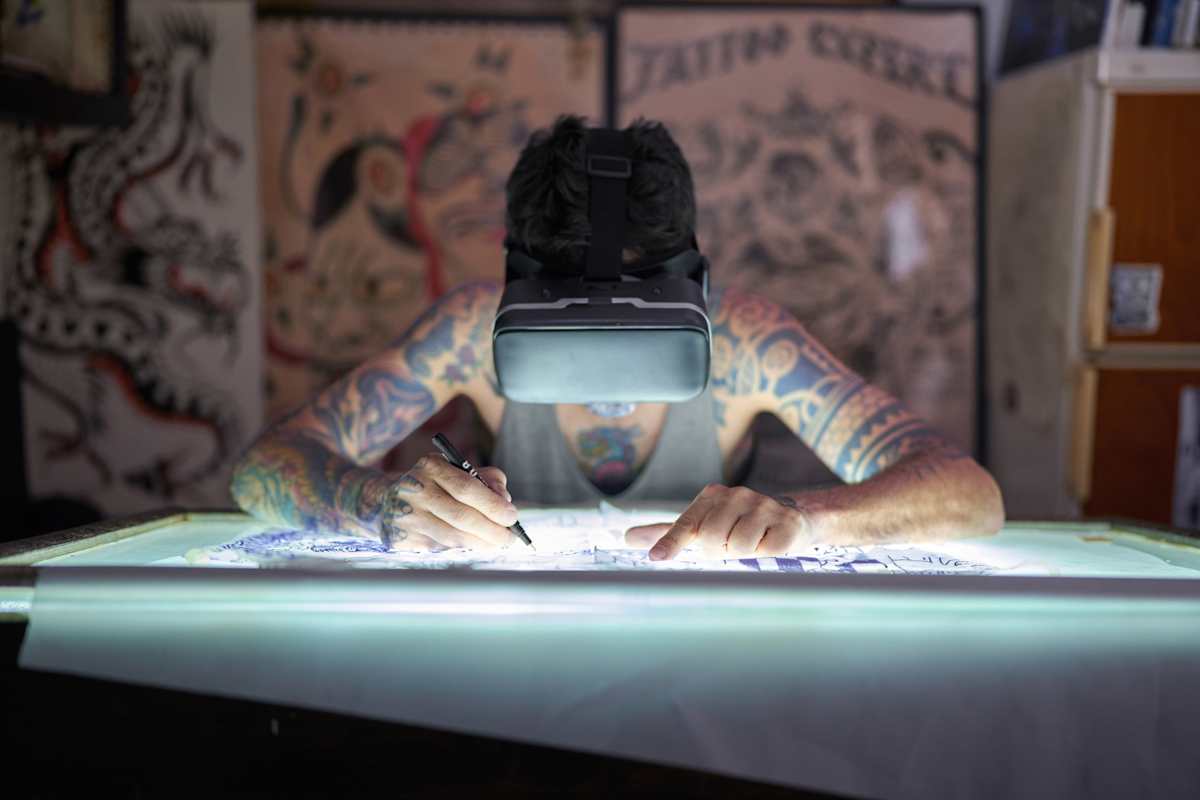 A male tattoo artist wearing a virtual reality headset in a studio hunched over a light table 