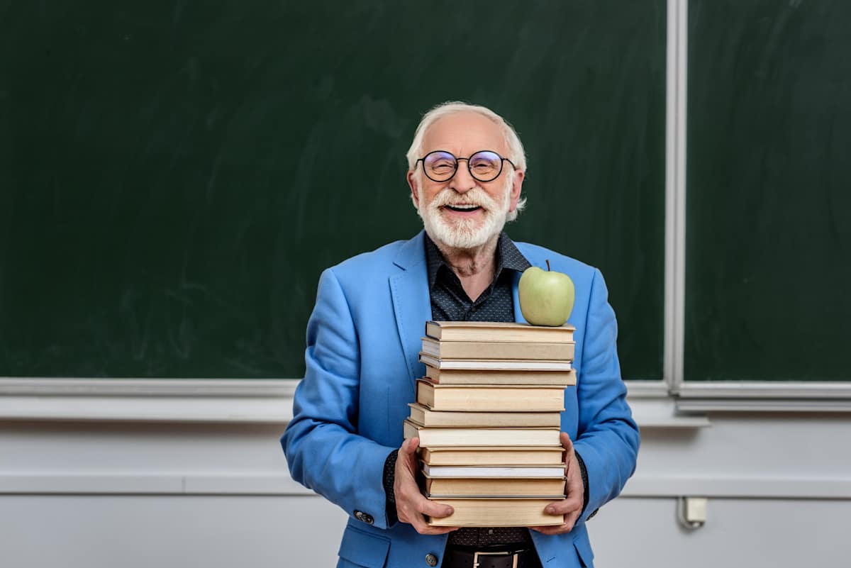 Grey-haired older man holding books in front of a blackboard.
