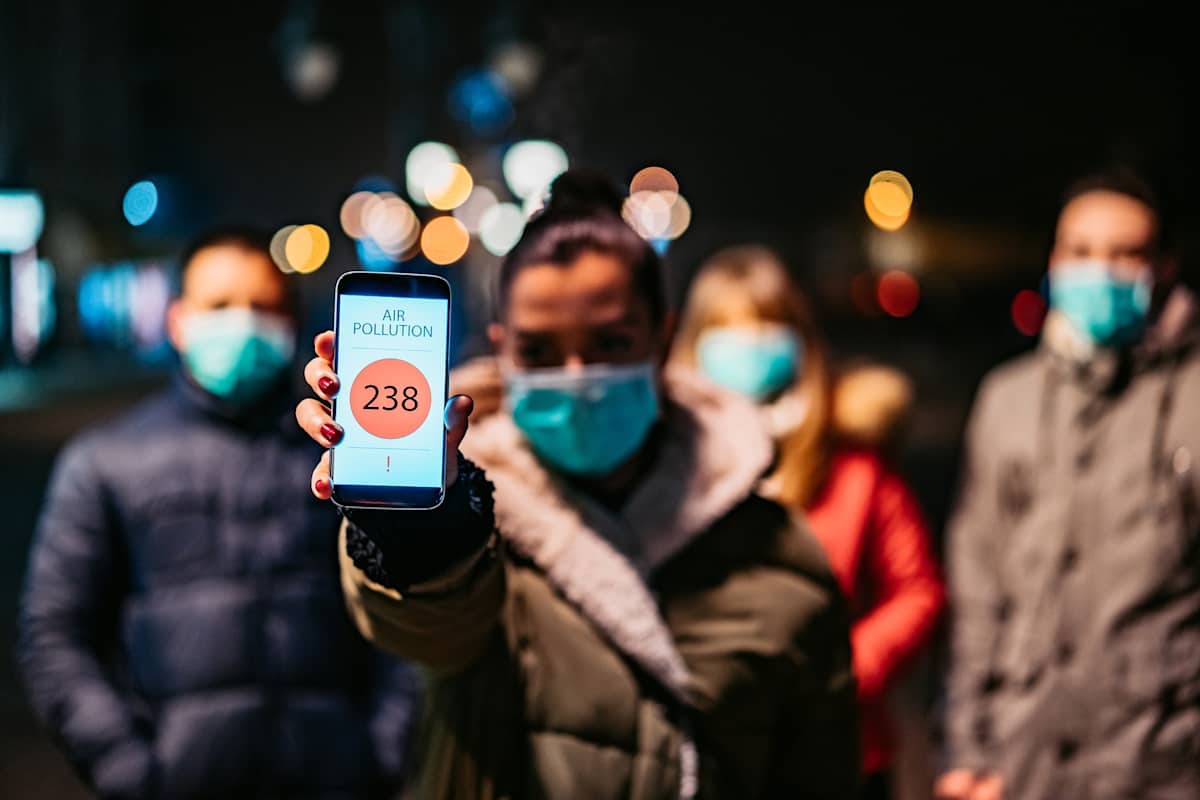 Young woman with pollution mask showing mobile pollution application in which is shown high air pollution in city.