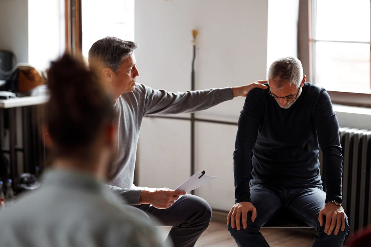 A support group session, with a seated man comforting another man with a hand on his shoulder