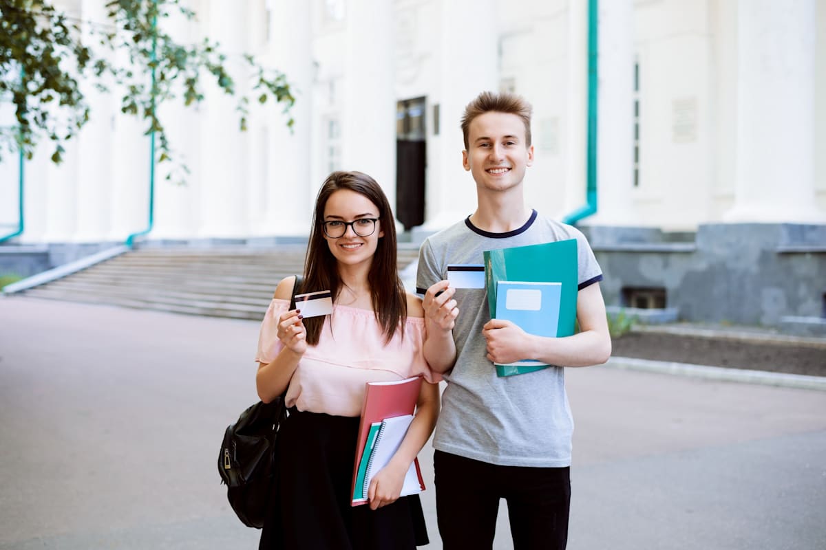 Two best friends smiling, holding credit cards and showing them to the camera in front of old university