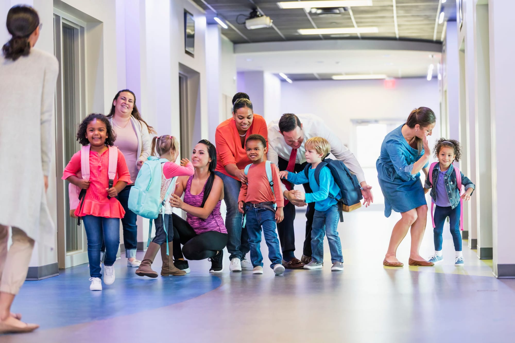 A multi-ethnic group of five preschool children, each with a parent, standing in a school hallway, getting words of encouragement.