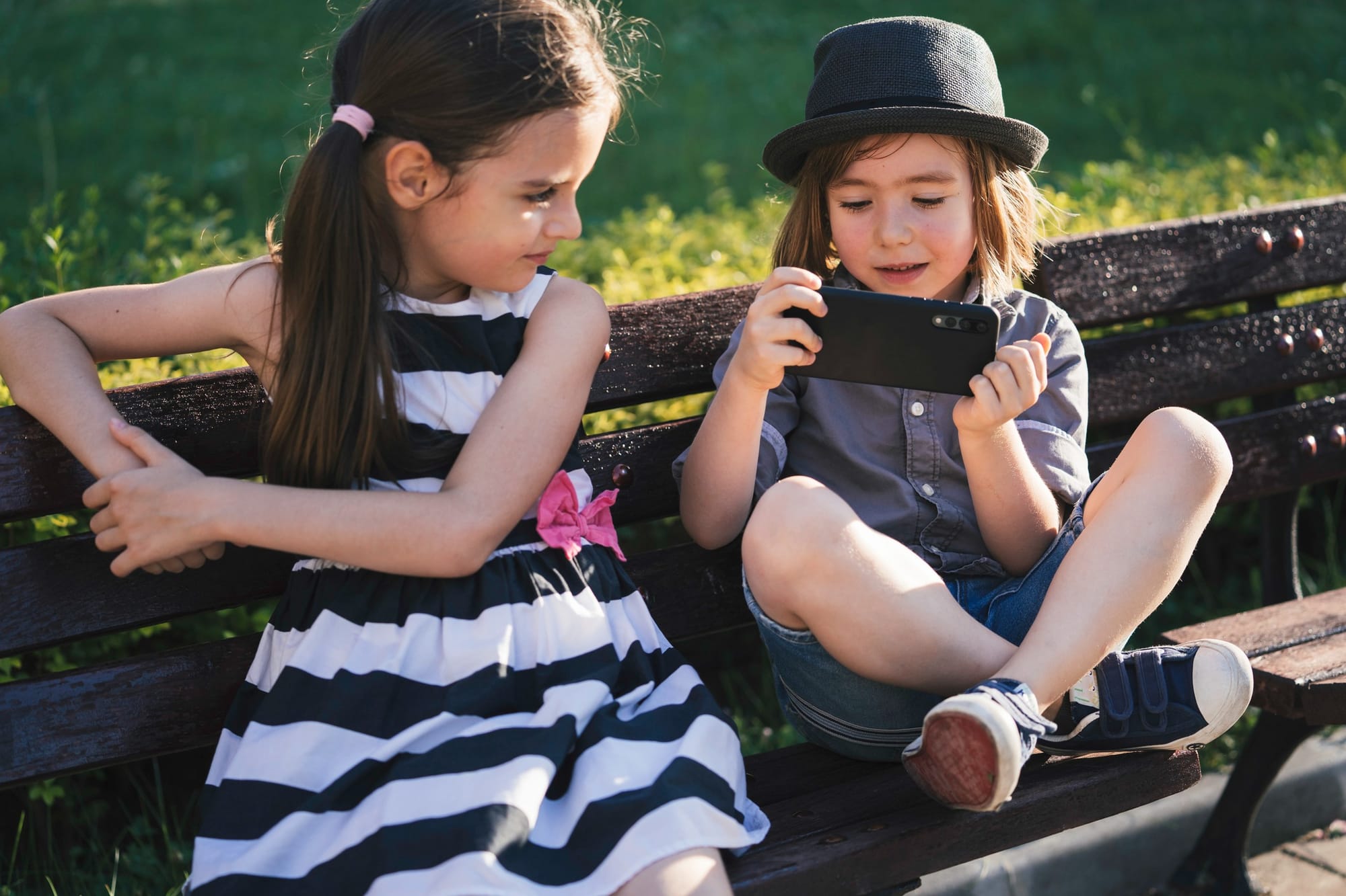 Two children sitting on a park bench, one playing on a mobile phone, the other not.