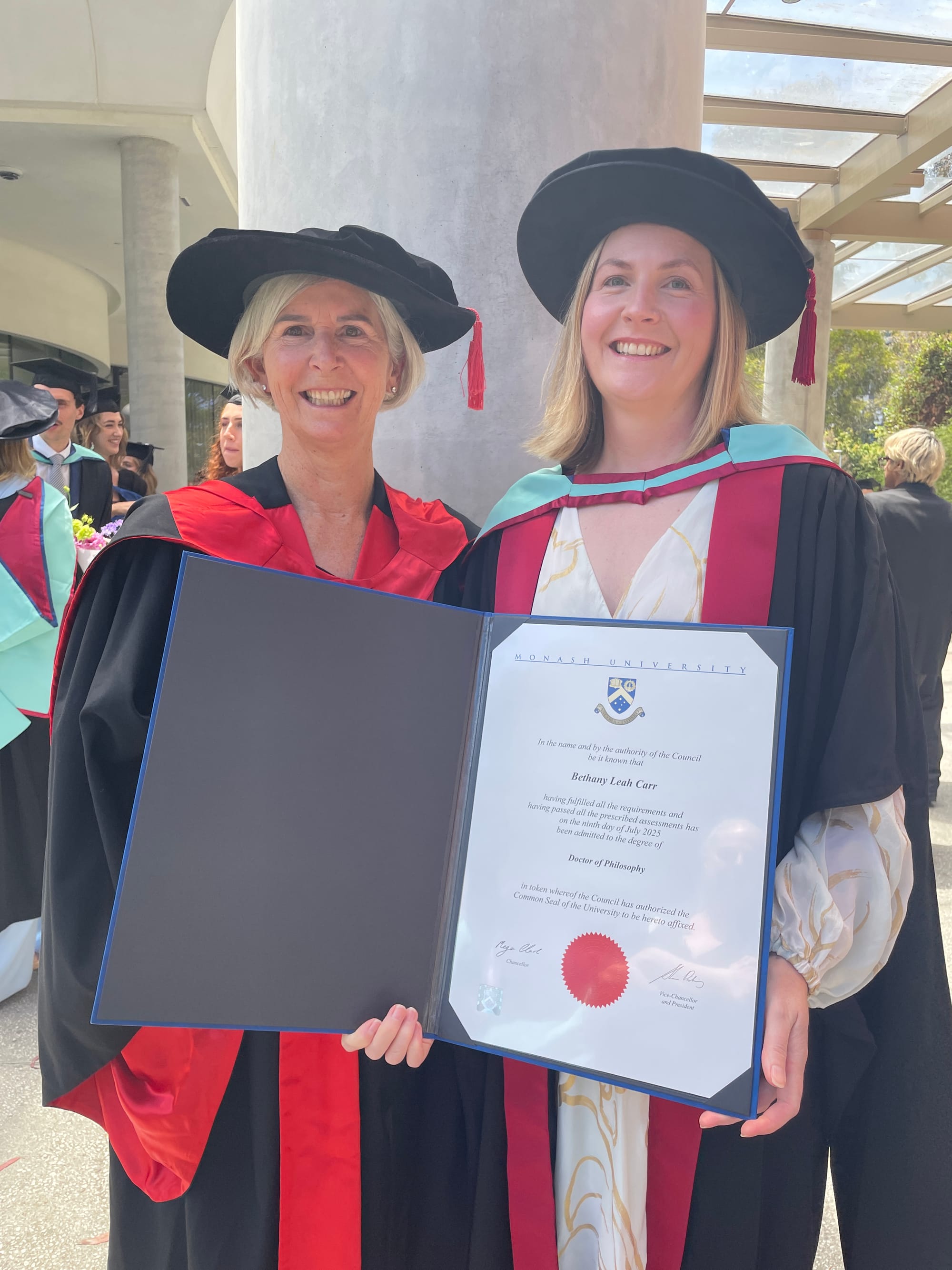 A smiling Jenny Gamble and Dr Bethany Carr in academic garb, Dr Carr holding her PhD document