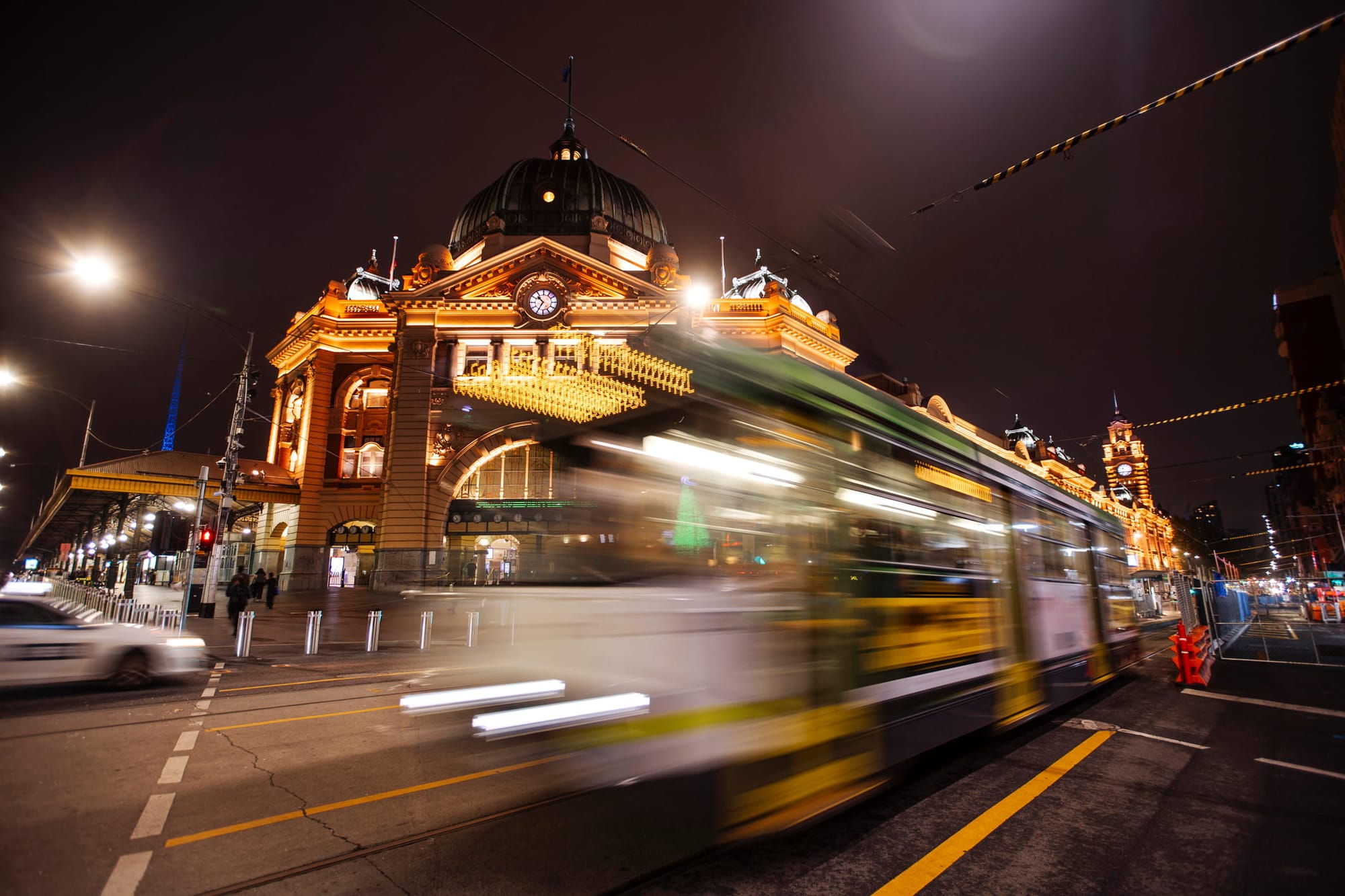 A vibrant night scene of an illuminated Flinders Street station as a tram blurs past in the foreground. 