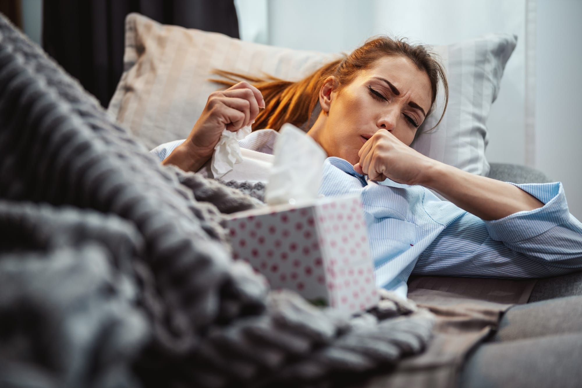 Young woman is lying sick at home couch with paper handkerchief in her hand, coughing.