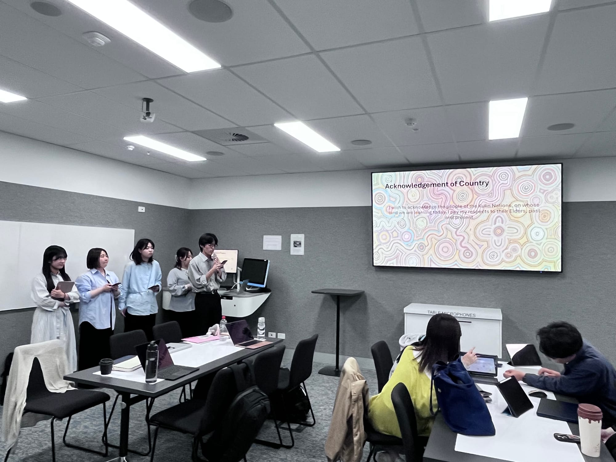 Japanese students standing with devices observing a screen with Acknowledgement of Country 