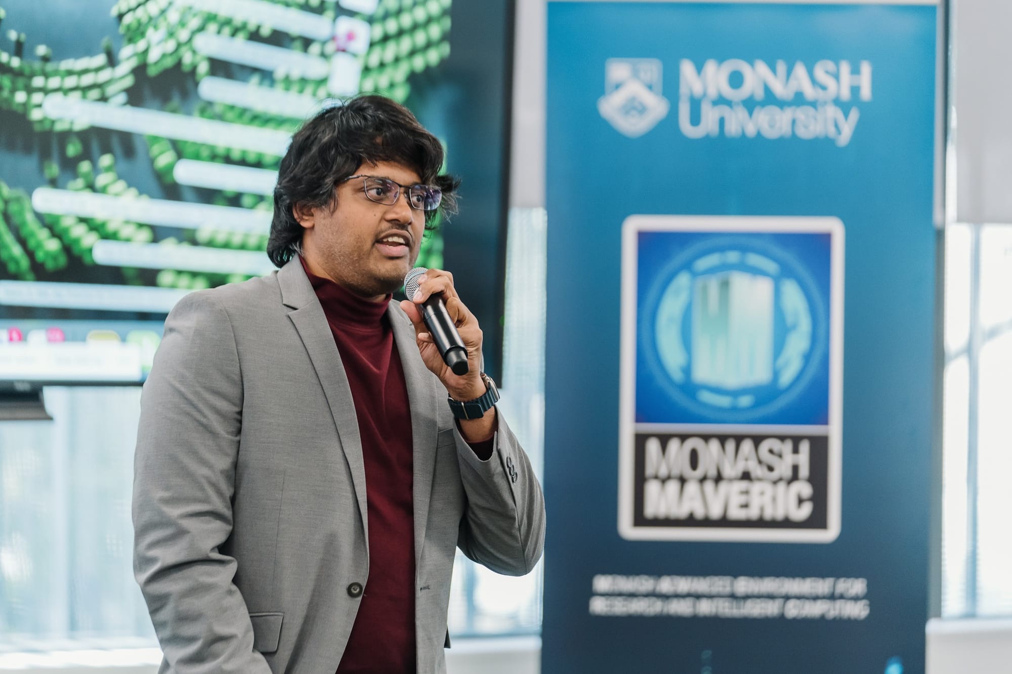 Man holding a microphone, speaking in front of a Monash University banner