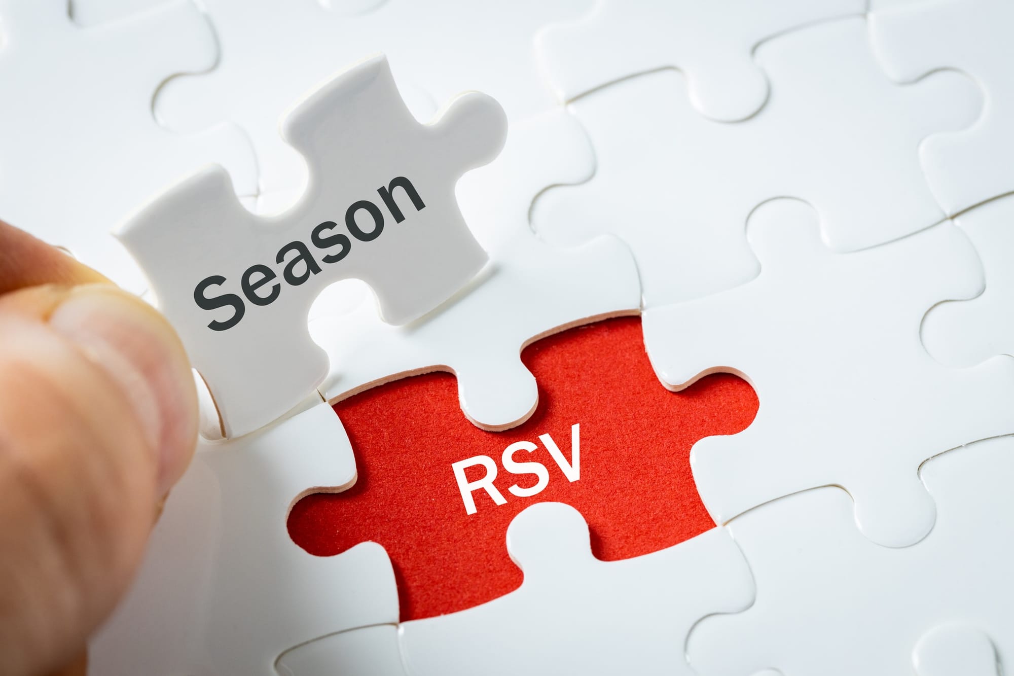 White jigsaw puzzle, one piece missing with "RSV" on a red background, and a person's hand holing a piece reading "Season"