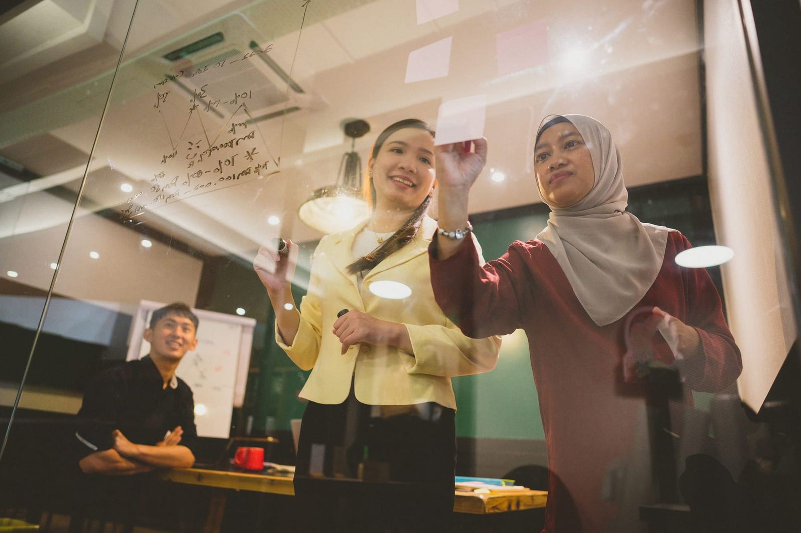 Two Asian women collaborating, writing on a clear glass wall
