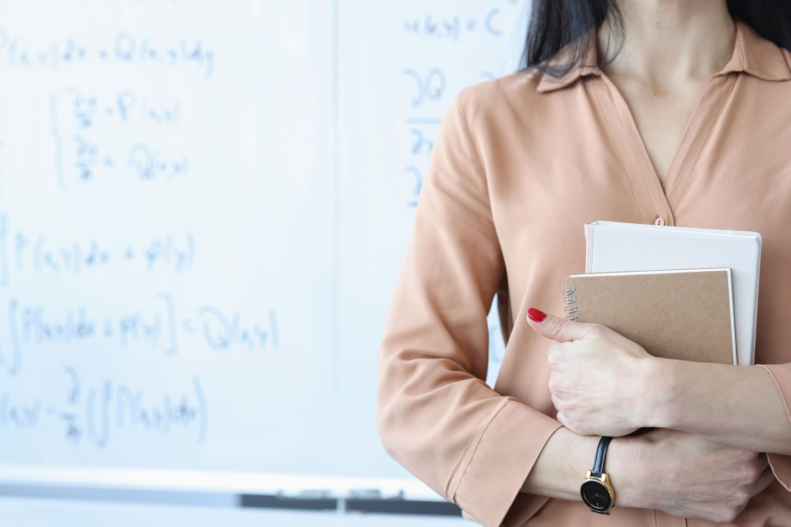 Female teacher holding books near whiteboard with blurred formulas written on it.