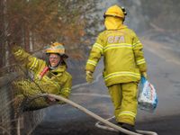 Two firefighters dressed in yellow CFA protective gear at a fence during a bushfire, one straining to climb through a fence