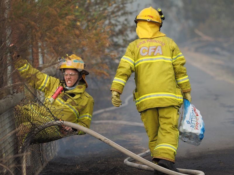 Two firefighters dressed in yellow CFA protective gear at a fence during a bushfire, one straining to climb through a fence