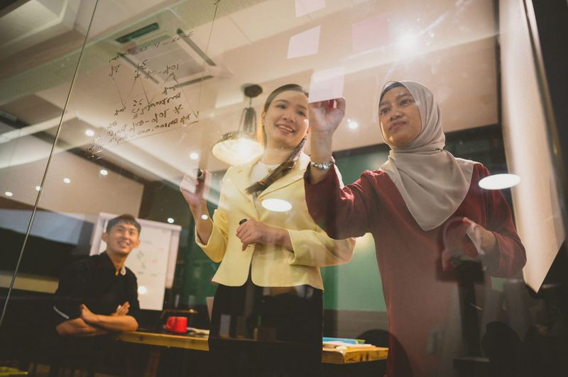 Two Asian women collaborating, writing on a clear glass wall