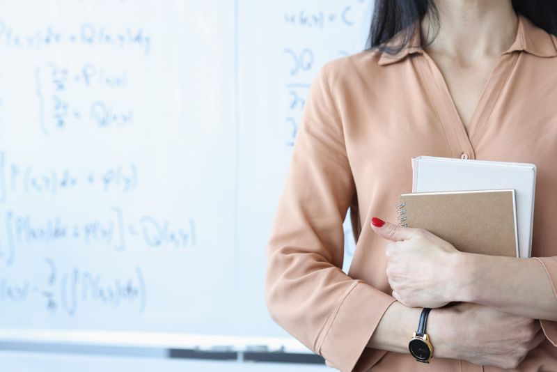 Female teacher holding books near whiteboard with blurred formulas written on it.