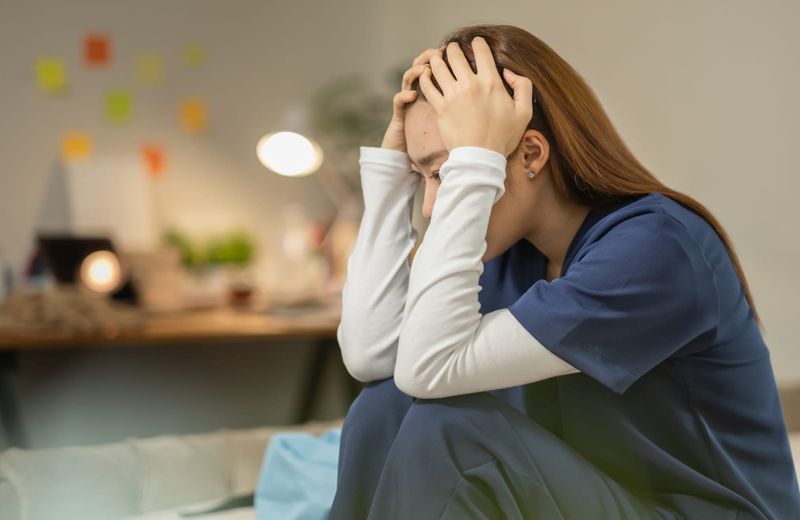 Asian woman in blue scrubs sitting with her head in her hands looking stressed