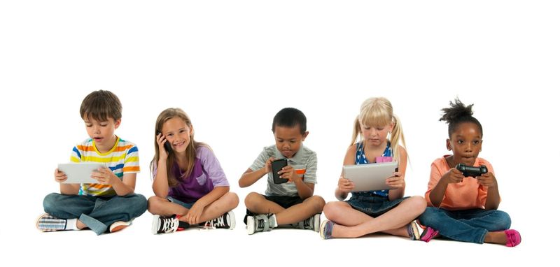 A diverse group of children sitting cross-legged with a variety of hand held devices.