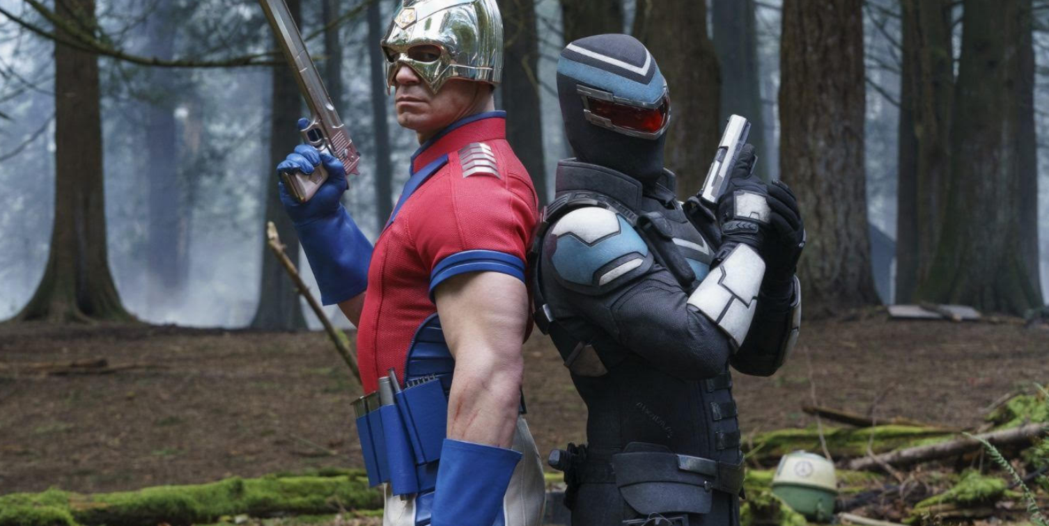 John Cena as Peacemaker and Freddie Stroma as Vigilante stand back-to-back with weapons drawn in a forest scene from the HBO Max series Peacemaker.