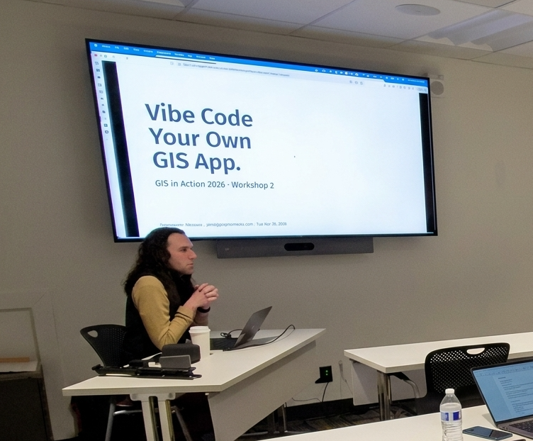 A presenter sits at a desk in a conference room, gesturing while teaching a workshop titled "Vibe Code Your Own GIS App" for the GIS in Action 2026 conference. The large wall-mounted screen displays the workshop title and subtitle, with a laptop open on the desk and attendee workstations visible in the foreground.