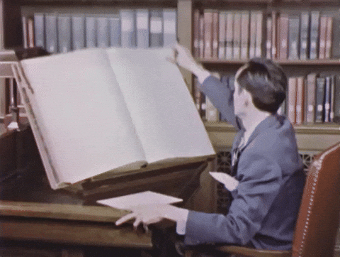 A person in a library sits at a large wooden desk, turning the oversized pages of an antique ledger or reference book, with rows of bookshelves visible in the background — illustrating the concept of doing traditional research homework before engaging with a contractor or AI.