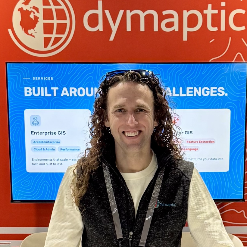 Christopher Moravec smiling at the dymaptic booth at Esri UC 2026, with a screen reading "built around your challenges" behind him.