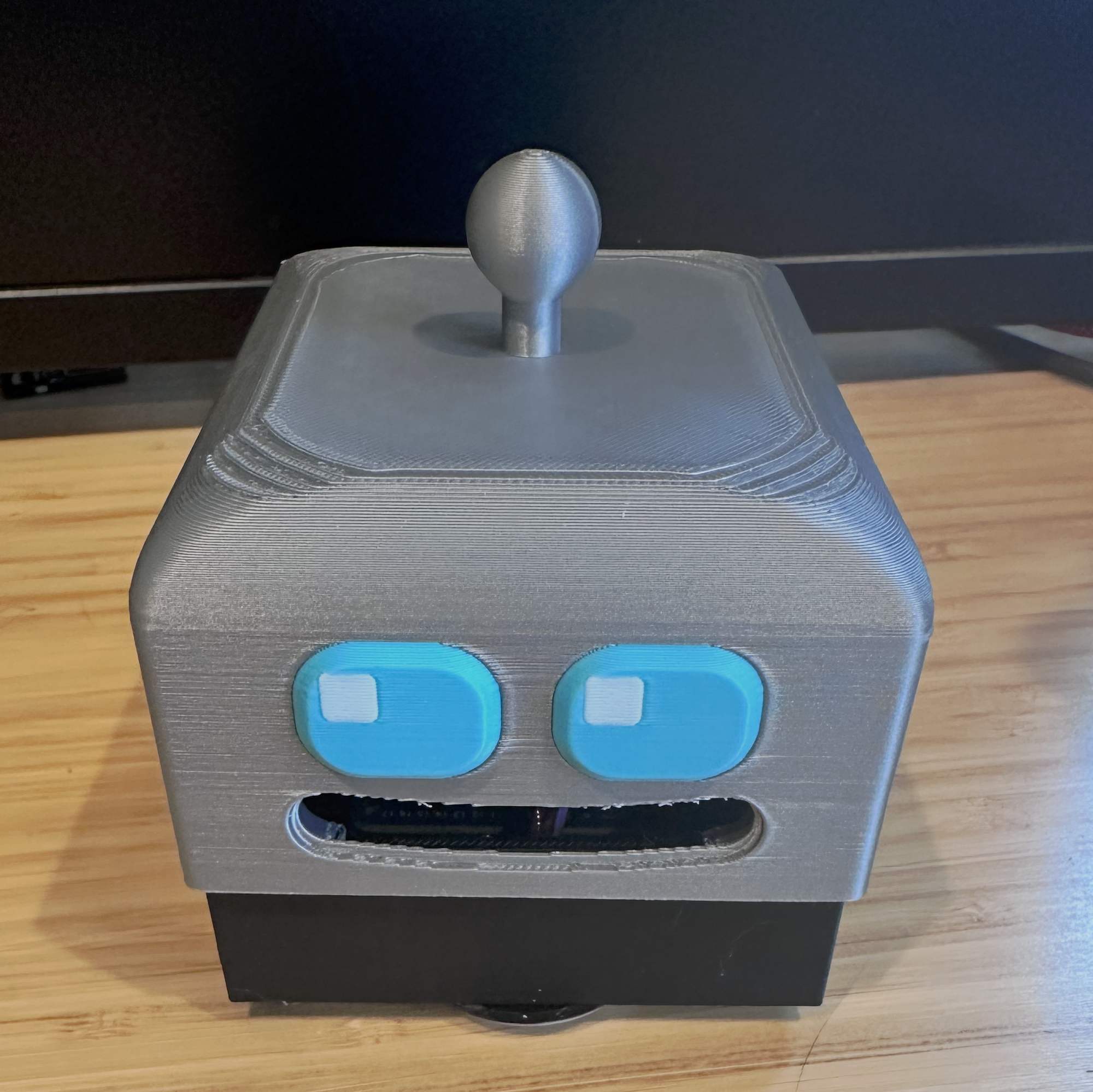 A 3D-printed gray robot head sitting on a wooden desk, featuring two bright blue rounded rectangular eyes, a slot-like mouth revealing internal electronics, and a small rounded antenna on top — the physical DeskBot described as being entirely designed by the AI Jaws.