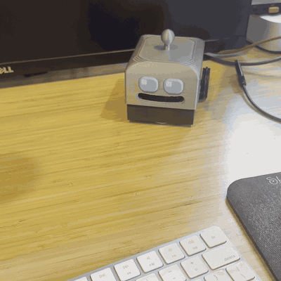 A small 3D-printed robot head sitting on a wooden desk near a keyboard and monitor, featuring two circular eyes, a rectangular mouth, and a joystick-style antenna on top — the physical DeskBot that was fully designed and engineered by the AI agent Jaws.
