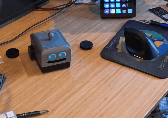 A small 3D-printed robot with a boxy gray body, two glowing teal eyes, and black wheels sits on a wooden desk beside a computer mouse and dances by spinning around and going back and forth a bit.