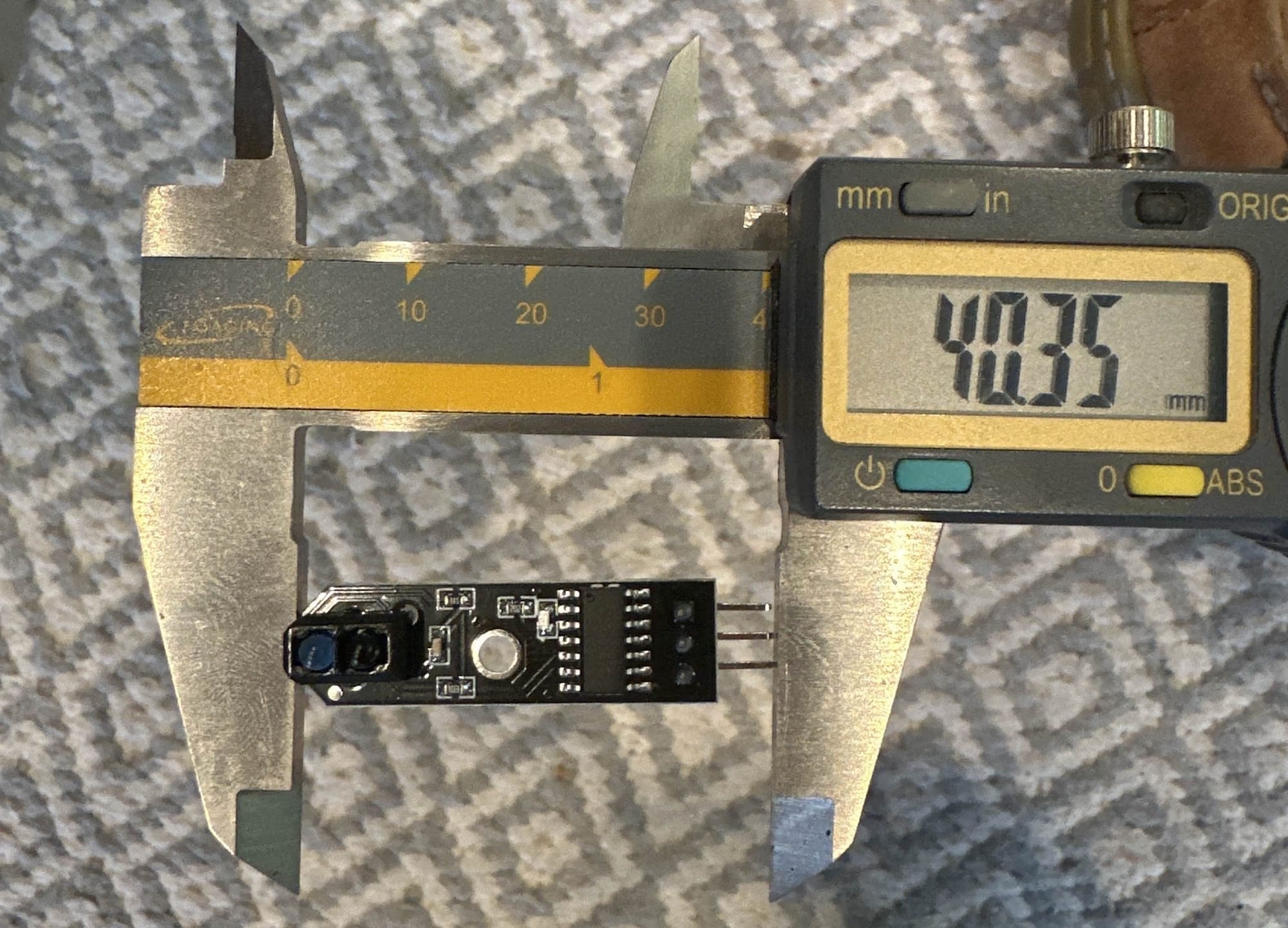 A small electronic sensor module being measured lengthwise with digital calipers, which display 40.35 mm, illustrating the challenge of relying on AI to interpret reference measurements from photos rather than reading them directly from the instrument.