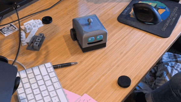 A small cube-shaped robot with two glowing teal eyes and wheels sits on a wooden desk, surrounded by a keyboard, sticky notes, and 3D-printed parts, slowly driving towards the edge and eventually pushing a small object off.