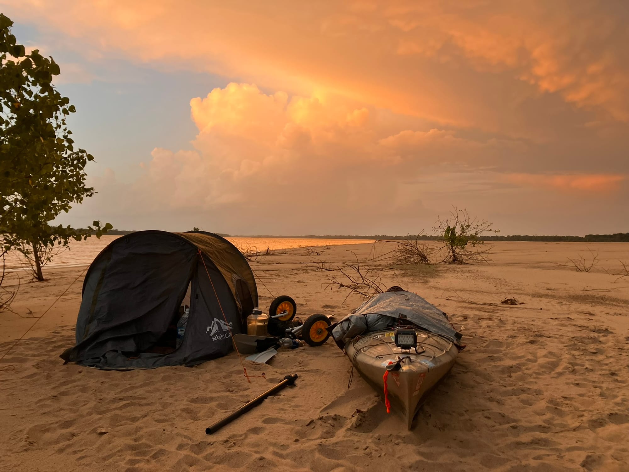A tent and a paddling boat rest on the sand as the sun sets.