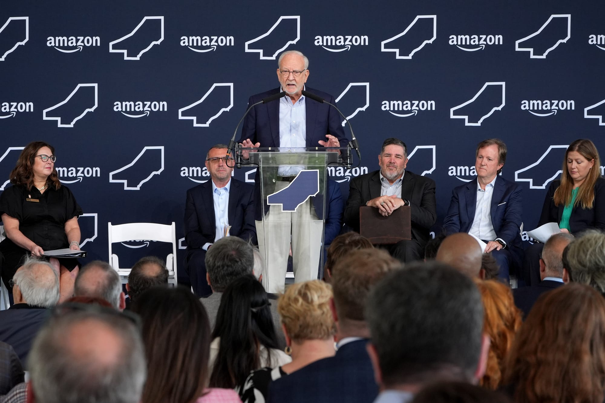 A white man in glasses and a dark jacket stands at a podium at a press conference. He stands in front of an Amazon logo-covered wall