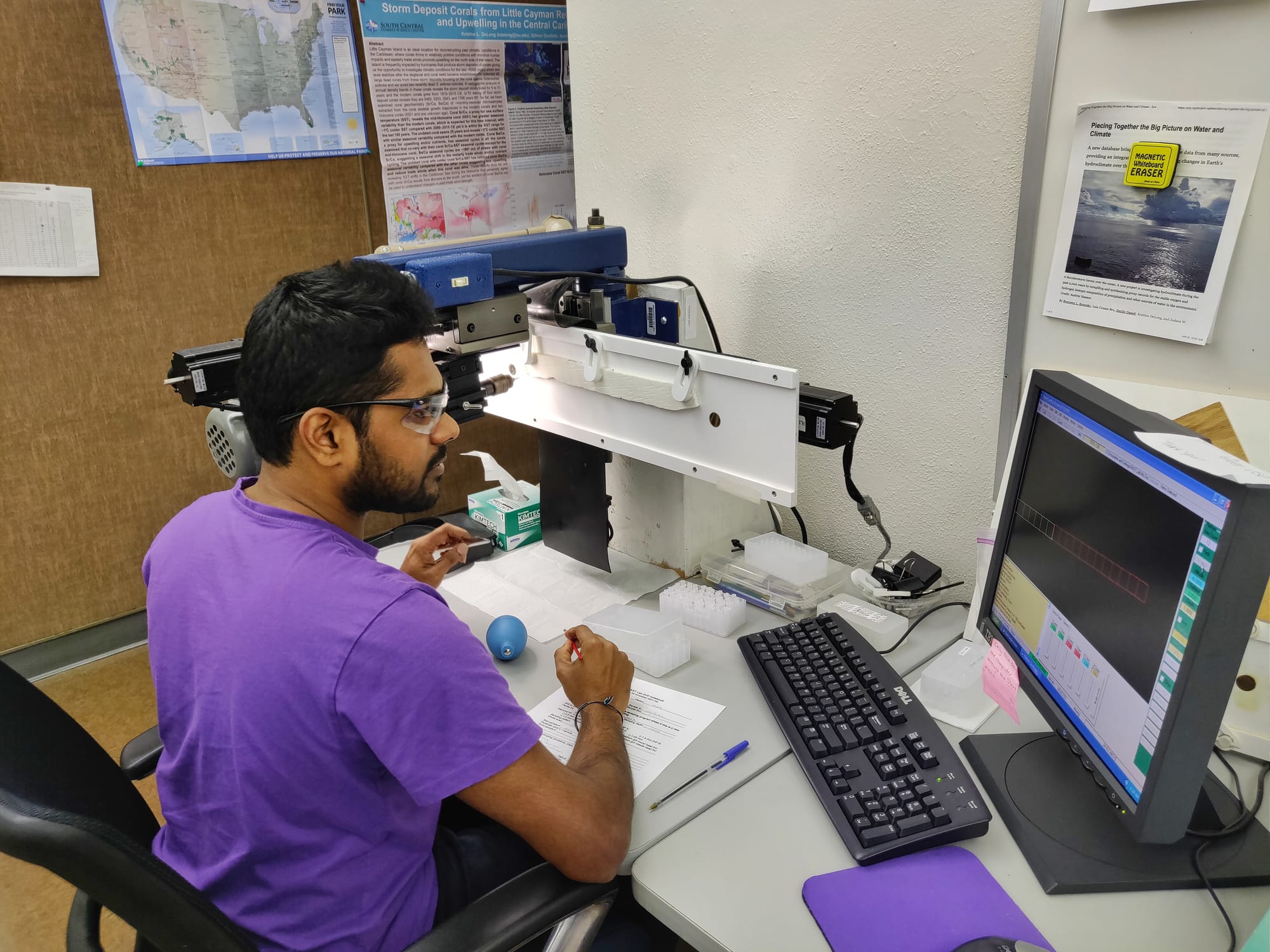 A grad student in lab glasses sits at a desk in front of equipment and looks at computer screen