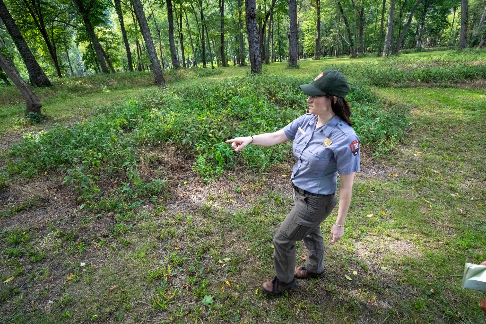 A woman in a green national parks hat and a short-sleeve button up adorned with patches and badges points to a grassy mound