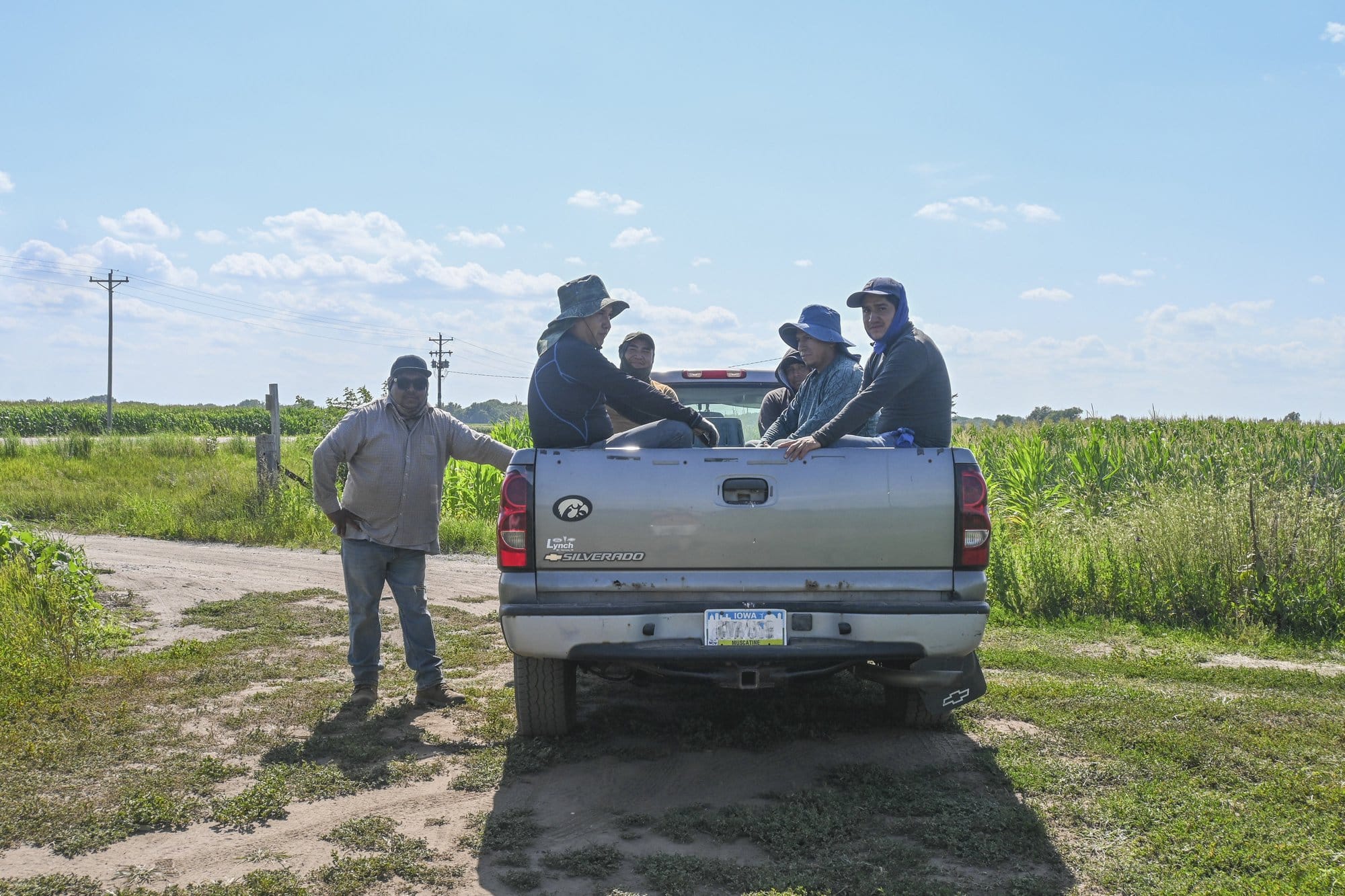 A team of farmworkers ride in the back of a truck with sun hats on, while one stands next to the drivers door