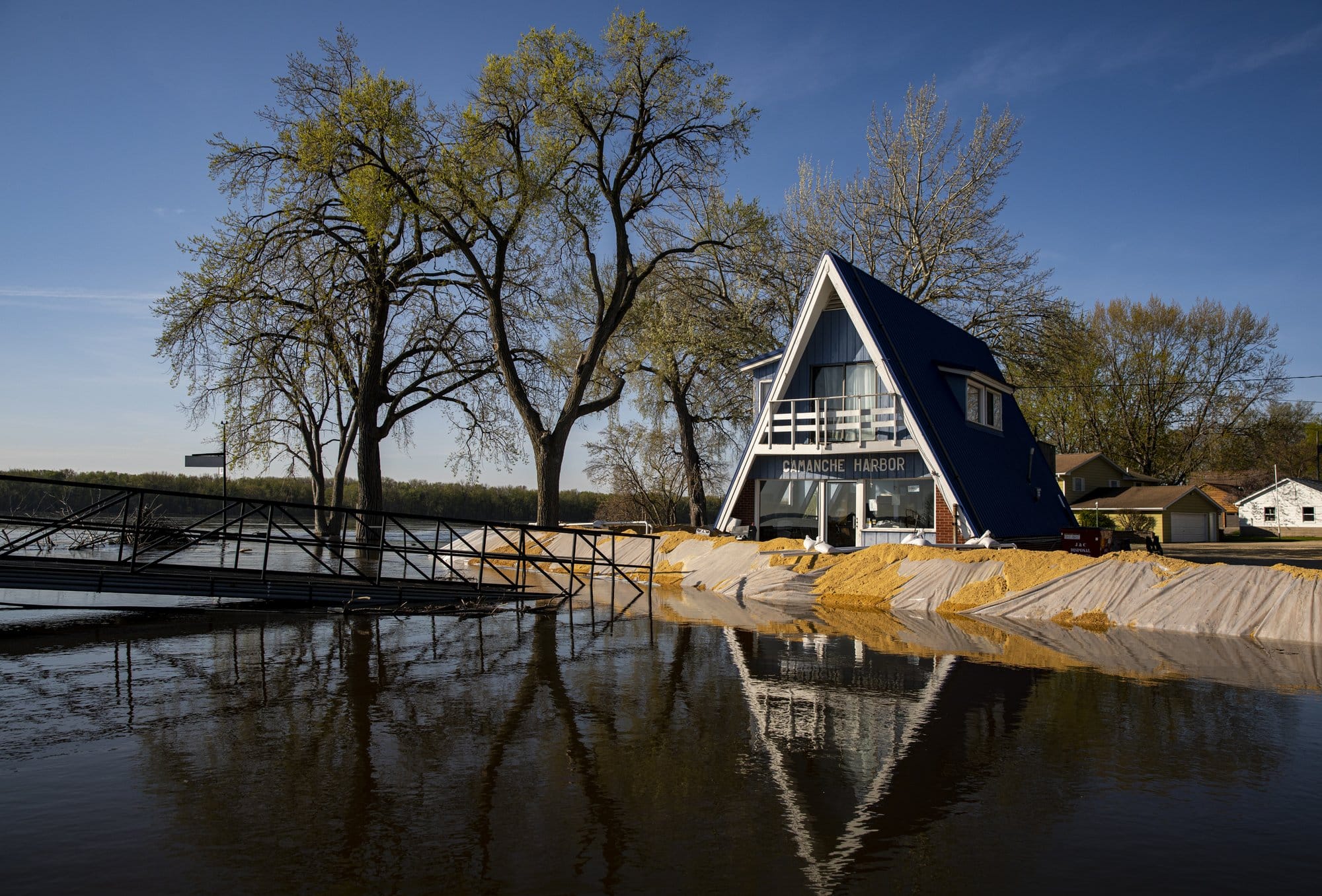 Sand weighs down a tarped barrier placed along the edge of a marina in front of a small, triangle-shaped harbor house