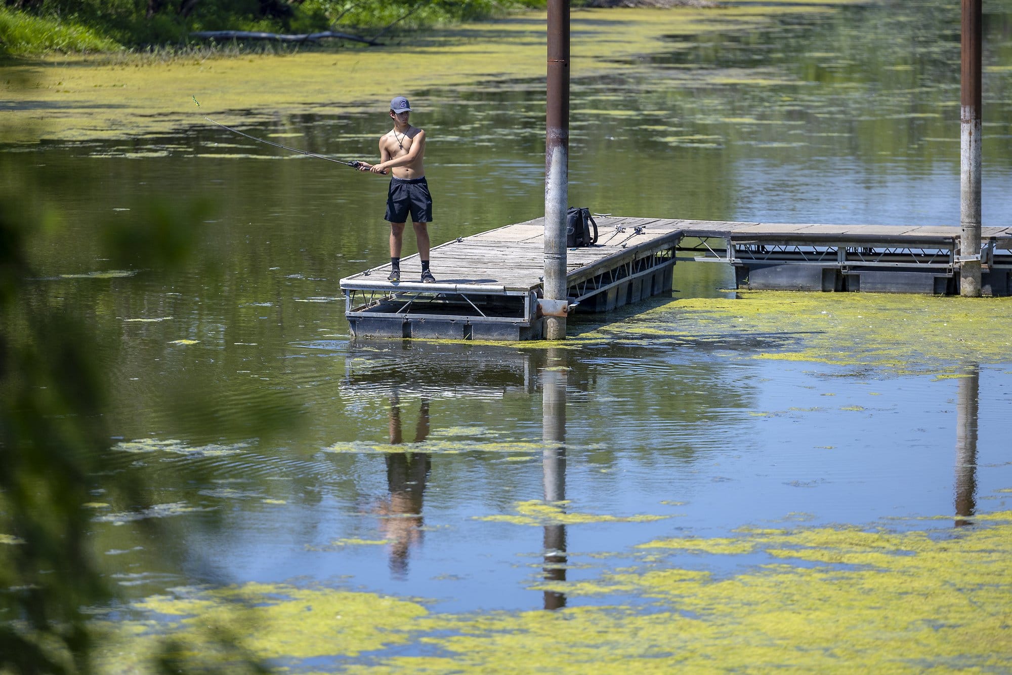 A man casts a fishing line off of a boat dock. Most of the water's surface is covered in a green film