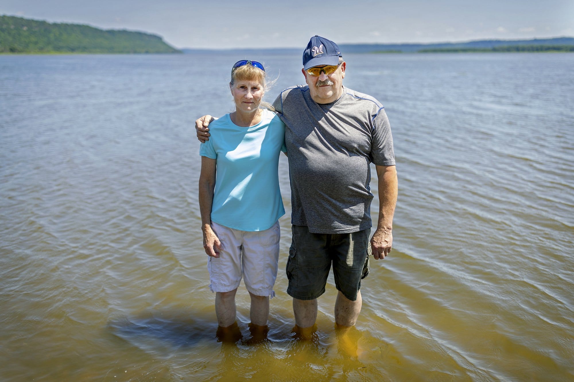 A man and woman stand in shallow water on a sunny day with their arms around eachother