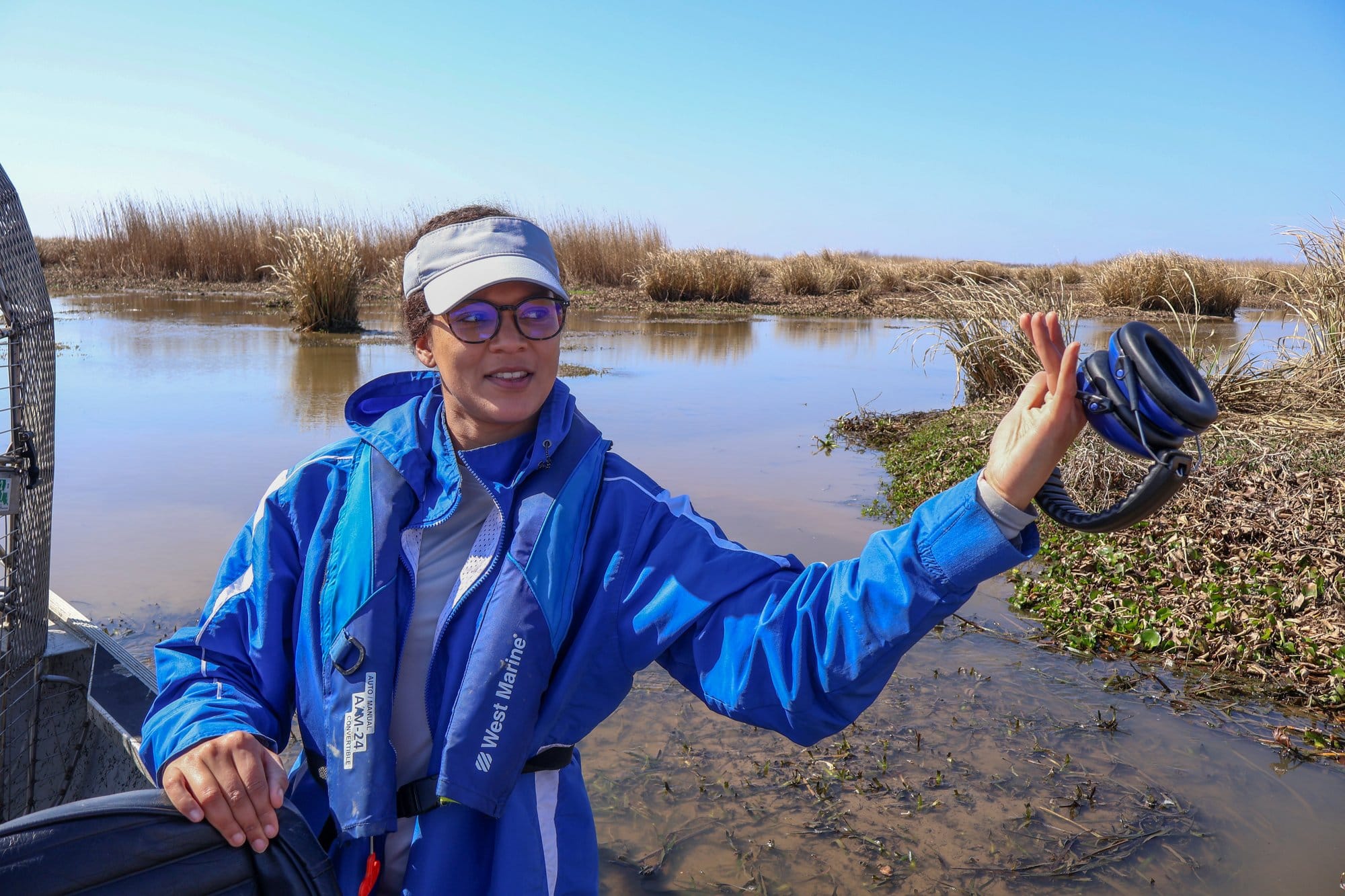 A woman in a blue waterproof coastal jacket and visor stands in a boat on the marsh