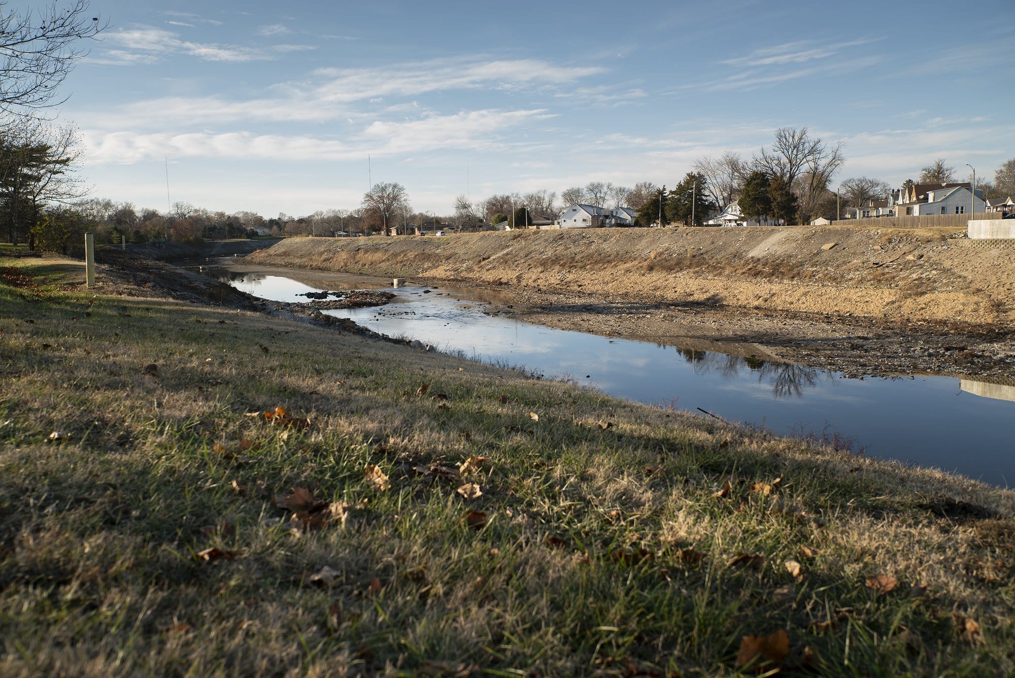 The River Des Peres drainage channel in south St. Louis, with shallow water running between grassy banks and homes visible in the background.