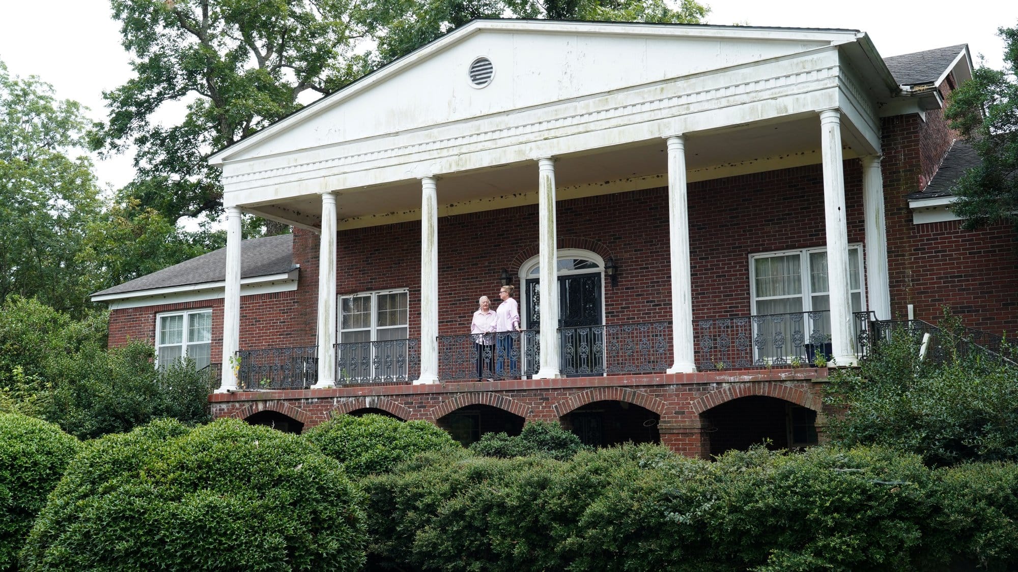 Two women stand on the deck of a brick house with white pillars