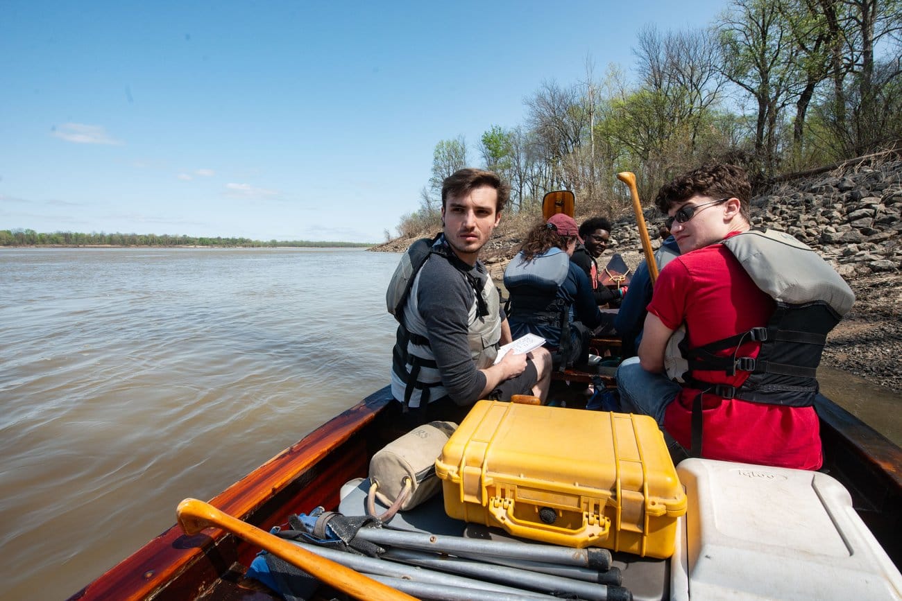 Two young men sit in a canoe near a riverside. There is gear behind them and two young women ahead of them.