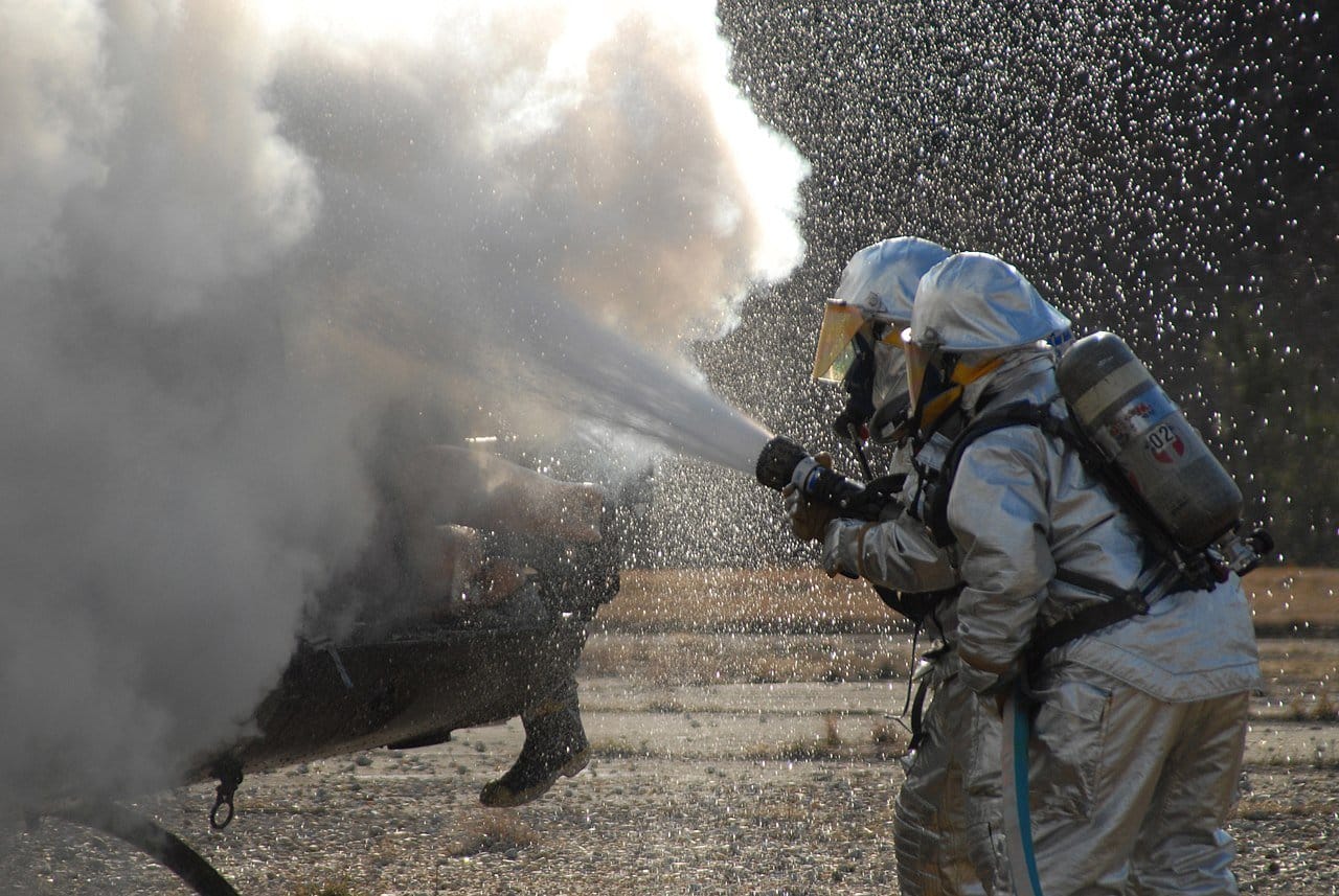 Two firefighters in protective gear spray foam onto a burning helicopter during a training exercise.
